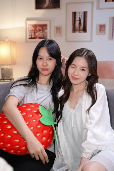 Two young women with long dark hair sit closely together on a couch, one holding a large red strawberry plush toy, while framed photos decorate the wall behind them. The cozy, playful scene reflects the warm and dreamy aesthetic of the "Ditto" series promotion.