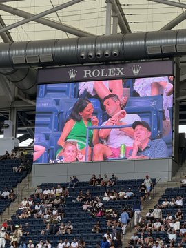 A crowd watches the US Open tennis tournament at Arthur Ashe Stadium, with fans visible on the large Rolex-branded jumbotron screen.