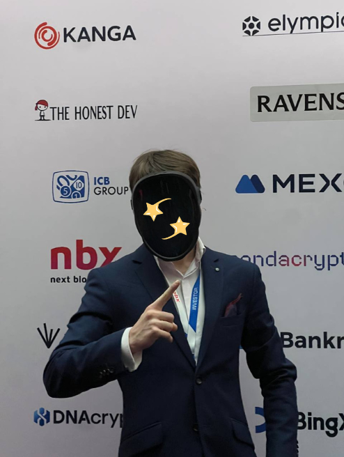 A person in a navy suit wearing a black mask with yellow stars poses in front of a sponsor backdrop featuring logos for Kanga, The Honest Dev, Ravens, MEXC, NBX, and Bankr.