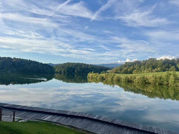 A scenic landscape photograph showing a calm lake reflecting a blue sky with wispy clouds, framed by forested hills and a wooden boardwalk in the foreground.