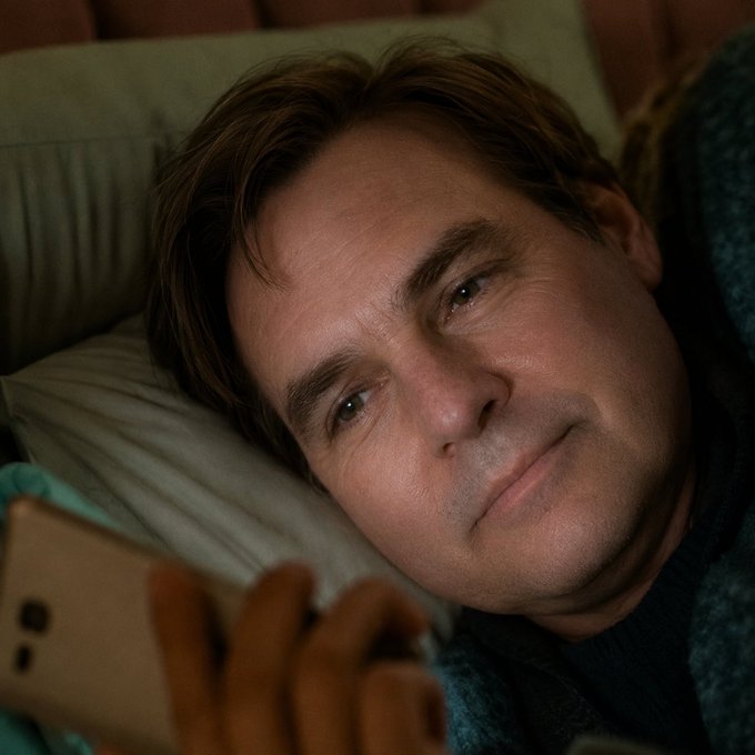A close-up shot of actor Jim Carrey lying in bed with a contemplative expression, looking at a smartphone with a gold-colored case.