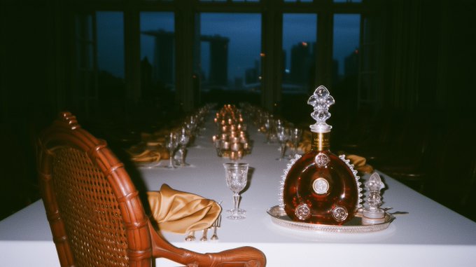 An opulent dining table setup featuring an ornate crystal decanter with amber liquid, crystal glassware, golden napkins, and a wicker chair, set against a dark city skyline backdrop.