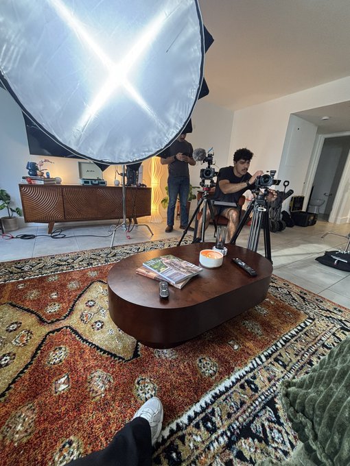 A film production setup in a living room shows a large lighting umbrella, a camera operator, and a boom mic operator, with a coffee table in the foreground.
