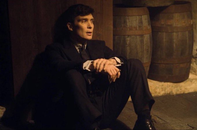 A young man in a dark suit sits on the floor leaning against a wooden wall next to wooden barrels in a dimly lit cellar, looking upward with an anxious expression.