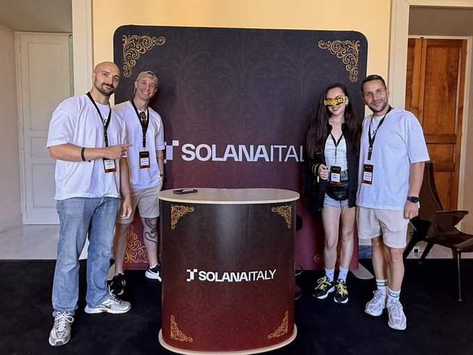 The image features a group of four men and two women posing for a photo next to a booth with a sign that says "Solana Italy". The people are standing around a large, circular display featuring the Solana logo. One person on the right side is wearing a yellow mask, which adds a fun and quirky element to the scene. The group appears to be enjoying themselves as they pose for the photo in front of the booth.