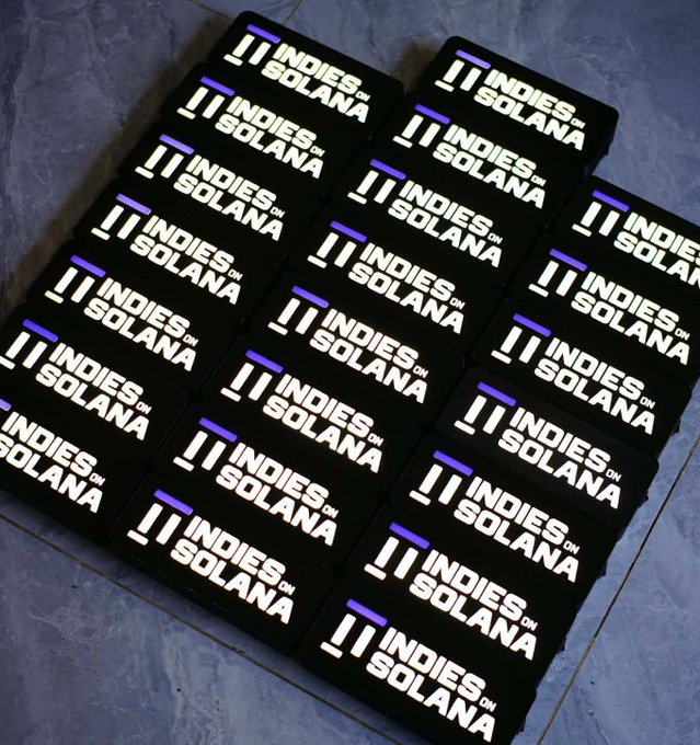 Multiple black t-shirts laid out in rows, each printed with the white text '!! INDIES ON SOLANA' along with a small purple accent line.