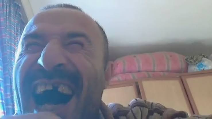 A close-up shot of a man with a goatee laughing enthusiastically, set against a simple background with a bed visible behind him.