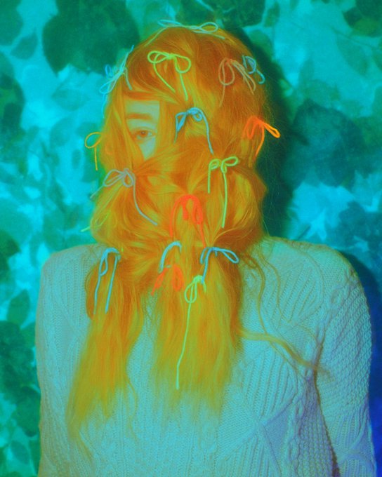 An artistic portrait of a person wearing a white sweater and a colorful knitted beard and headpiece made of orange and neon-colored yarn, with a teal-tinted background.