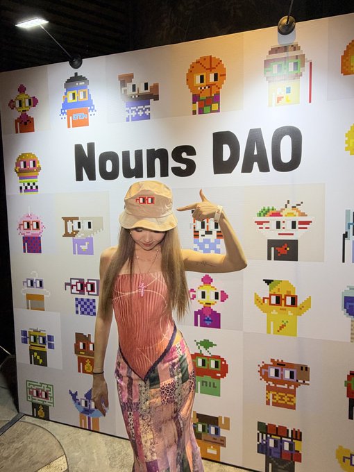 A woman in a bucket hat, pink top, and floral pants poses in front of a "Nouns DAO" wall display featuring various pixel art character designs.