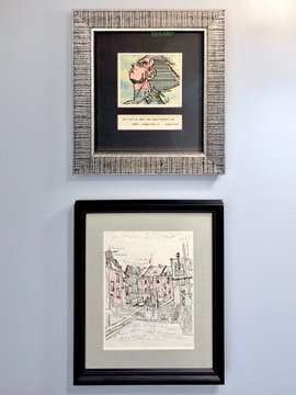 Two framed artworks hanging on a light blue wall: the upper one contains a small figurine with a pink parasol against a textured background, while the lower one displays a black-and-white sketch of a castle or chateau.