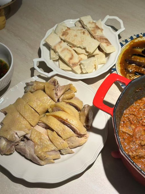 A meal spread featuring a plate of sliced poached chicken, a dish of flatbread pieces, a red pot of stewed tomato-based curry, and another pot of darker sauce on the side.