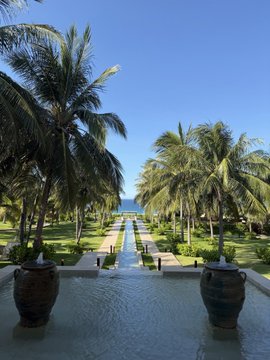 The image features a beautiful tropical garden with palm trees and a pool area. The pool has two large vases placed at opposite ends, adding an artistic touch to the scene. In front of the pool, there are two potted plants that contribute to the overall aesthetic appeal of the space. The sky above is clear blue, creating a serene atmosphere for relaxation and enjoyment.