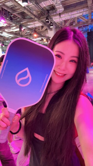 A smiling young woman with long dark hair holds up a purple paddle-shaped fan featuring a leaf icon at what appears to be a convention or event space.