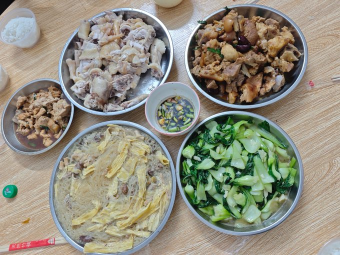 A spread of home-cooked Chinese dishes on a wooden table, including boiled pork pieces, braised pork trotters, egg and vermicelli soup, stir-fried bok choy, and a small dish of dipping sauce with garlic and chili.
