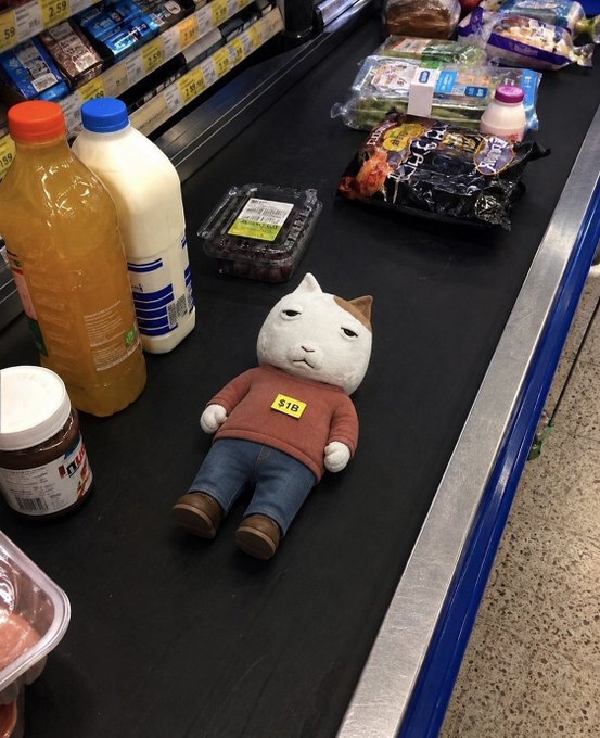 A plush cat toy wearing a red sweater with a '$15' sticker lies on a supermarket checkout conveyor belt alongside various grocery items including orange juice, milk, and snacks.