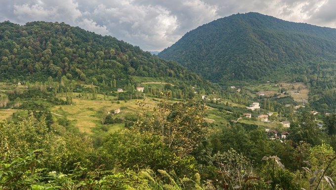 A scenic landscape view of lush green forested mountains and rolling hills with scattered houses under a cloudy sky.