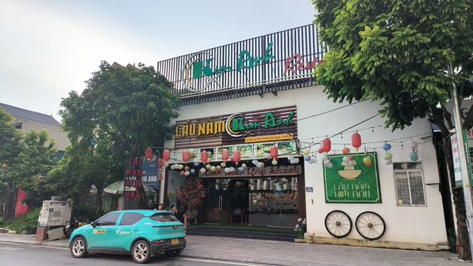 A street view shows a Vietnamese restaurant named 'Lẩu Nam Chú Rể' decorated with colorful lanterns and bicycles as decor, with a turquoise VinFast taxi parked in front.