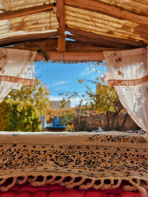 A view from inside a tent or covered seating area looking out over an ornate patterned rug toward a sunny garden with trees and a blue sky.