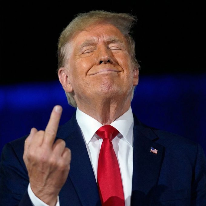 A portrait of Donald Trump in a dark suit and red tie, smiling with his eyes closed and raising his middle finger.