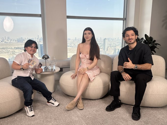 Three people posing on rounded white chairs in a high-rise apartment with a panoramic city skyline view, two holding Pokemon cards.