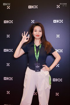 A young woman wearing a green "OKX" lanyard and a navy polo shirt makes an "OK" hand gesture while posing in front of an OKX-branded backdrop, tying in to the post's announcement of her presence at Token2049 in Singapore.