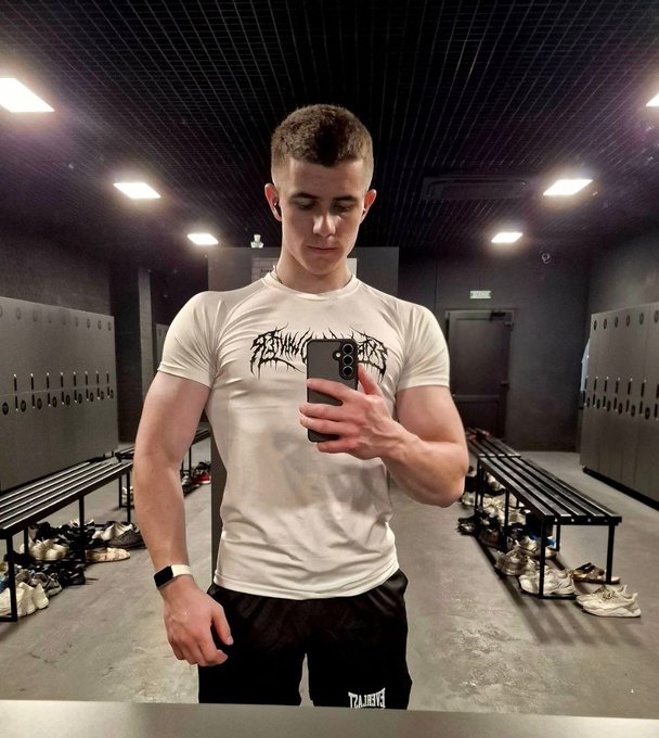 A young man taking a mirror selfie in a locker room, wearing a white t-shirt with a graphic design and black shorts, while holding a phone.