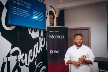 The image features an architect standing in front of a wall with a large screen displaying information about the "Meetup" event, which seems to be related to architecture or design. The architect appears to be giving a presentation or speech at this event. In addition to the main subject, there is also a person holding a cup and a clock visible in the scene.