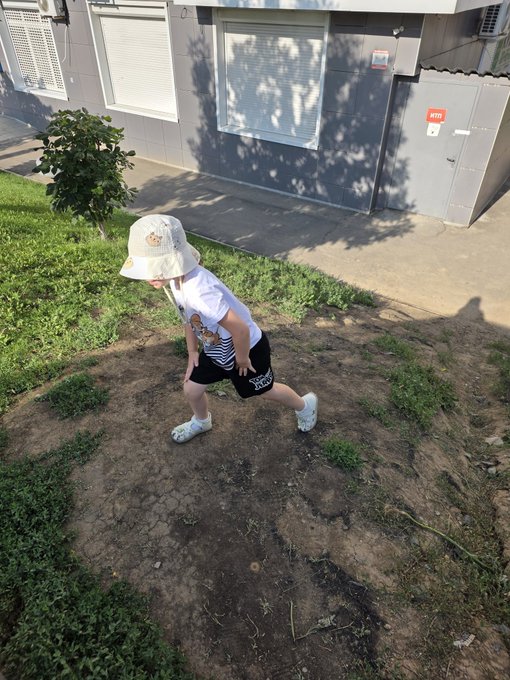 A young child wearing a beige bucket hat, white shirt, and black shorts walking across a grassy and dirt yard in front of a gray building.