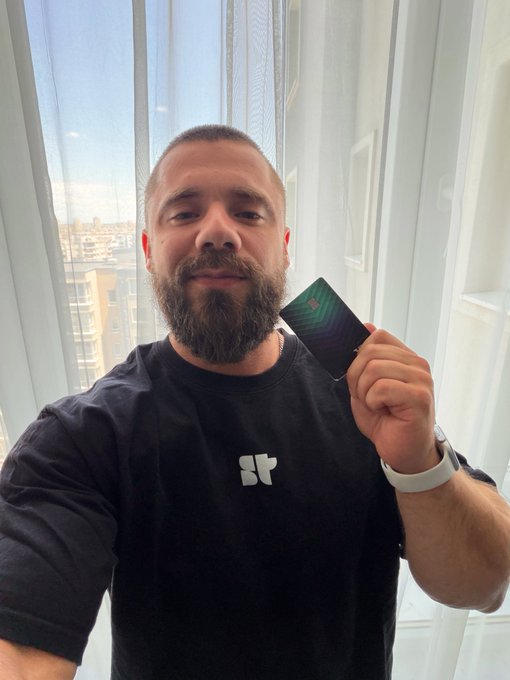 A bearded man in a black t-shirt holds up a dark credit/debit card while standing in front of a window, seemingly responding to critics by sharing his positive personal experience using the card for crypto-related daily expenses over the past two years.