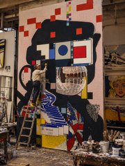 An artist wearing a mask works on a colorful, large-scale street art mural featuring a stylized black figure with red, white, and blue geometric shapes against a pink background.