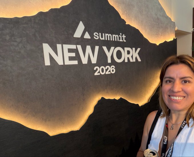 A smiling woman with brown hair standing in front of a Summit New York 2026 backdrop designed to look like a mountain landscape.