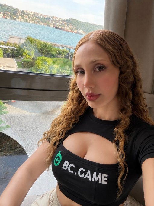 A woman with long wavy hair wearing a black crop top with the 'BC.GAME' logo takes a selfie by a window overlooking a scenic body of water and a ship.