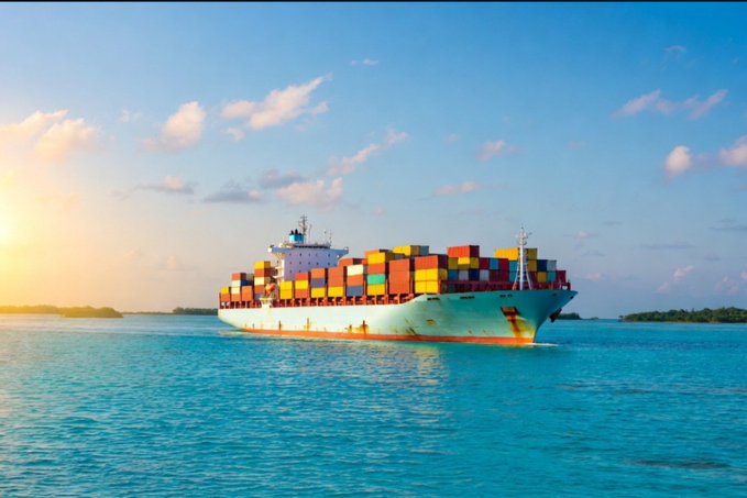 A large cargo ship loaded with colorful shipping containers sails across turquoise ocean waters at sunset.