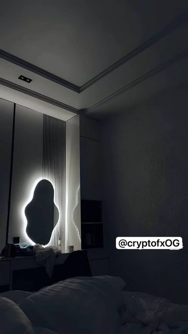 A dimly lit bedroom with a backlit mirror shaped like an irregular blob, with the watermark '@cryptofxOG' visible on the wall.
