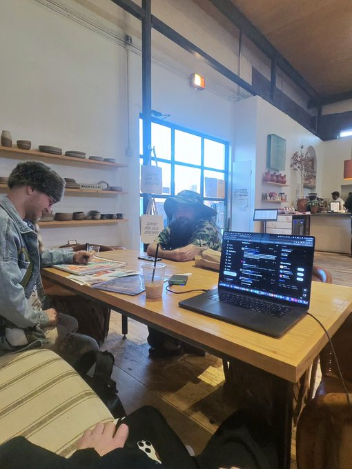 People sit around a wooden table in a cafe or shop, with a laptop showing dark-themed code or data in the foreground and shelves of bowls behind them.