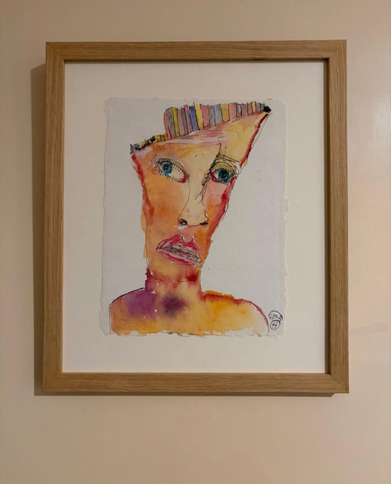 A framed watercolor painting of a stylized face with colorful striped hair, hanging on a beige wall.