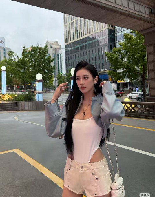 A young woman with long dark hair poses on a city street wearing a white top, light blue cropped jacket, and cream-colored shorts, with a small handbag.
