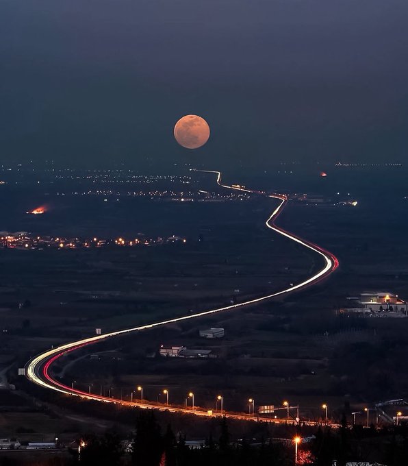 A winding highway glows with streaks of light from passing vehicles, leading the eye toward a large, orange full moon rising on the horizon, perfectly illustrating the "Way to moon" caption by transforming the illuminated road into a path toward the lunar destination.