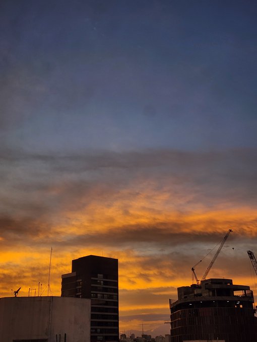A dramatic sunset paints the sky with vivid orange and blue hues above a city skyline featuring buildings under construction, complete with cranes silhouetted against the colorful clouds. The image's sweeping dusk atmosphere captures the contemplative mood of the morning greeting ("Bom dia!") and the cautionary note that Bitcoin hasn't yet hit its bottom ("lembre-se de que o #bitcoin ainda não chegou ao fundo fundo"), symbolizing the calm before a potential financial storm.