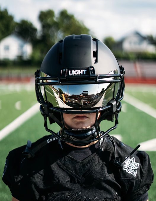 A close-up portrait of a football player wearing a black helmet with 'LIGHT' on the front and a mirrored visor that reflects a football field, standing on a green turf field.