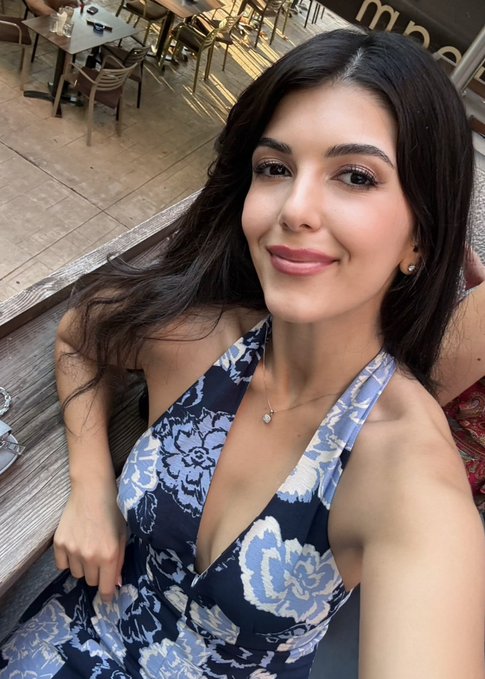 Selfie of a smiling woman with long dark hair wearing a blue and white floral halter top, seated at an outdoor cafe.