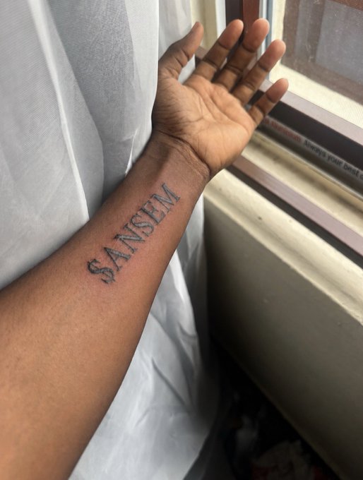 A person holds up their forearm near a window, displaying a bold black tattoo of the word "SANSEM" on their inner wrist. The image illustrates the post's theme of devotion to the ANSEM meme coin, showing a fan who took the unconventional route of getting the name permanently inked rather than purchasing the token.