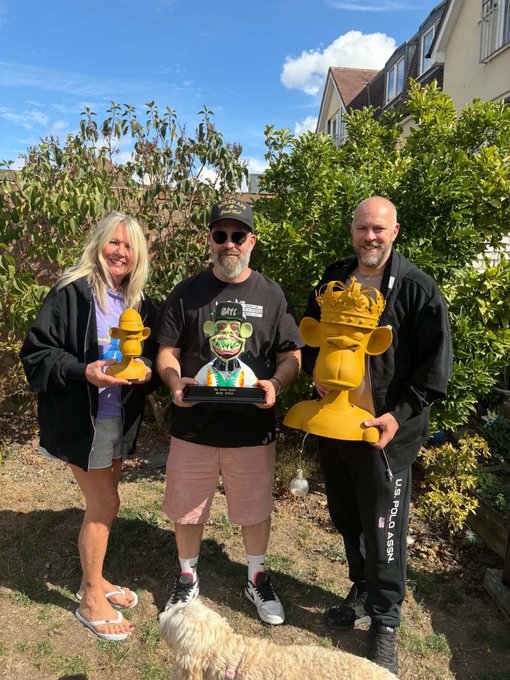 Three people posing outdoors with golden trophies; the woman on the left holds a small golden bear figurine, the man in the middle holds a trophy featuring a green character, and the man on the right holds a larger golden bear bust wearing a crown, with a fluffy white dog in the foreground.