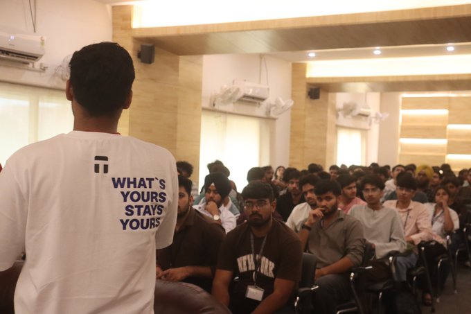 A speaker in a white t-shirt with "WHAT'S YOURS STAYS YOURS" printed on it addresses a packed auditorium of attentive attendees in India. The speaker faces a diverse crowd of young adults seated in rows, suggesting a well-attended talk or seminar.