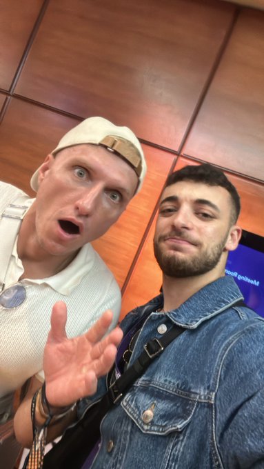 Two men pose for a selfie in what appears to be a conference or event space, with one wearing a white cap and white shirt making an exaggerated surprised expression while gesturing with his hand, and the other in a denim jacket offering a more composed look. A sign in the background partially reads "passed ground," adding to the casual, fun vibe that matches the "GM" (Good Morning) greeting.