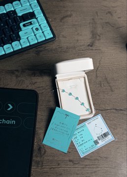 A small piece of jewelry with green beads is shown in its open white case with teal product tags, placed on a wooden desk next to a keyboard and a black mat.