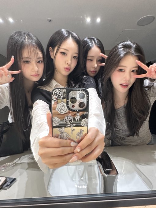 Four young women taking a mirror selfie, with the person in front holding a smartphone featuring a black case decorated with white roses.