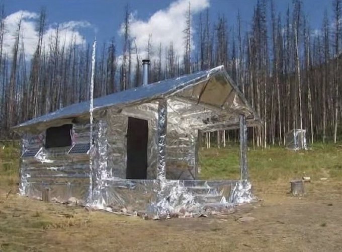 A small makeshift cabin or shelter covered entirely in reflective silver foil/insulation material sits in a clearing with burnt/dead trees in the background, featuring a metal chimney pipe and small windows.