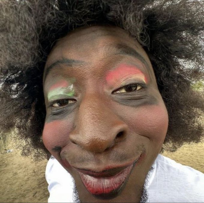 A close-up of a person with dark curly hair wearing colorful clown-style makeup with green, pink, and black accents around the eyes and cheeks.