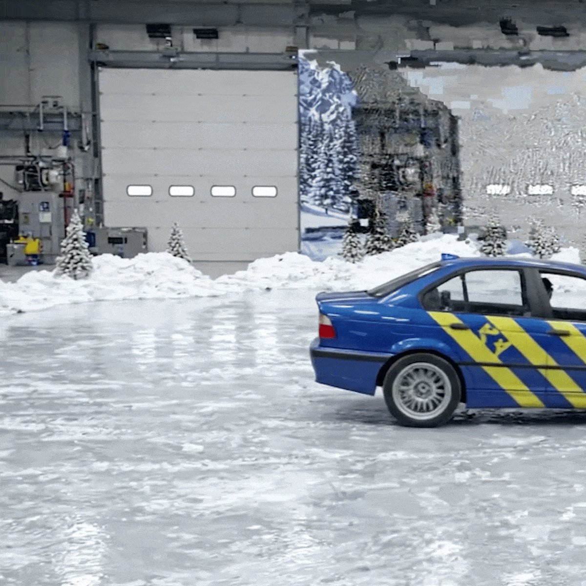 The image shows a blue car driving through an indoor testing area with snow-covered ground, which appears to be a garage or warehouse setting. The car is positioned near the center of the scene, moving towards the right side. There are also several pine trees visible in the background, adding a touch of nature to the industrial environment. The image seems to be related to an auction or event taking place at the testing area, as indicated by the caption "TESTING_/ALPINE1” auction ends in 2 hours".